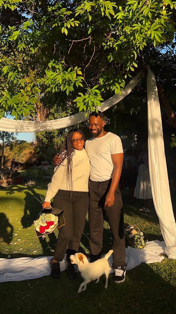 Gladman and Vongayi arm in arm in golden evening light beneath a garden tree draped in white fabric, their small white dog standing on the grass before them