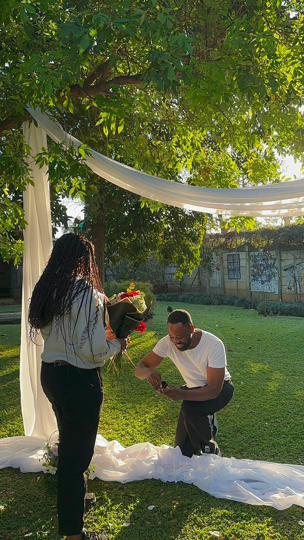 Gladman down on one knee holding the ring box while Vongayi, carrying a bouquet, stands beneath a draped garden arch